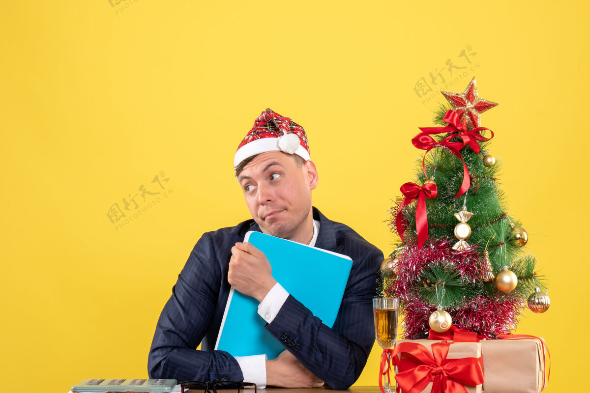 男前视图的商人紧紧地拿着他的文件坐在圣诞树附近的桌子上 黄色的礼物黄色人男人