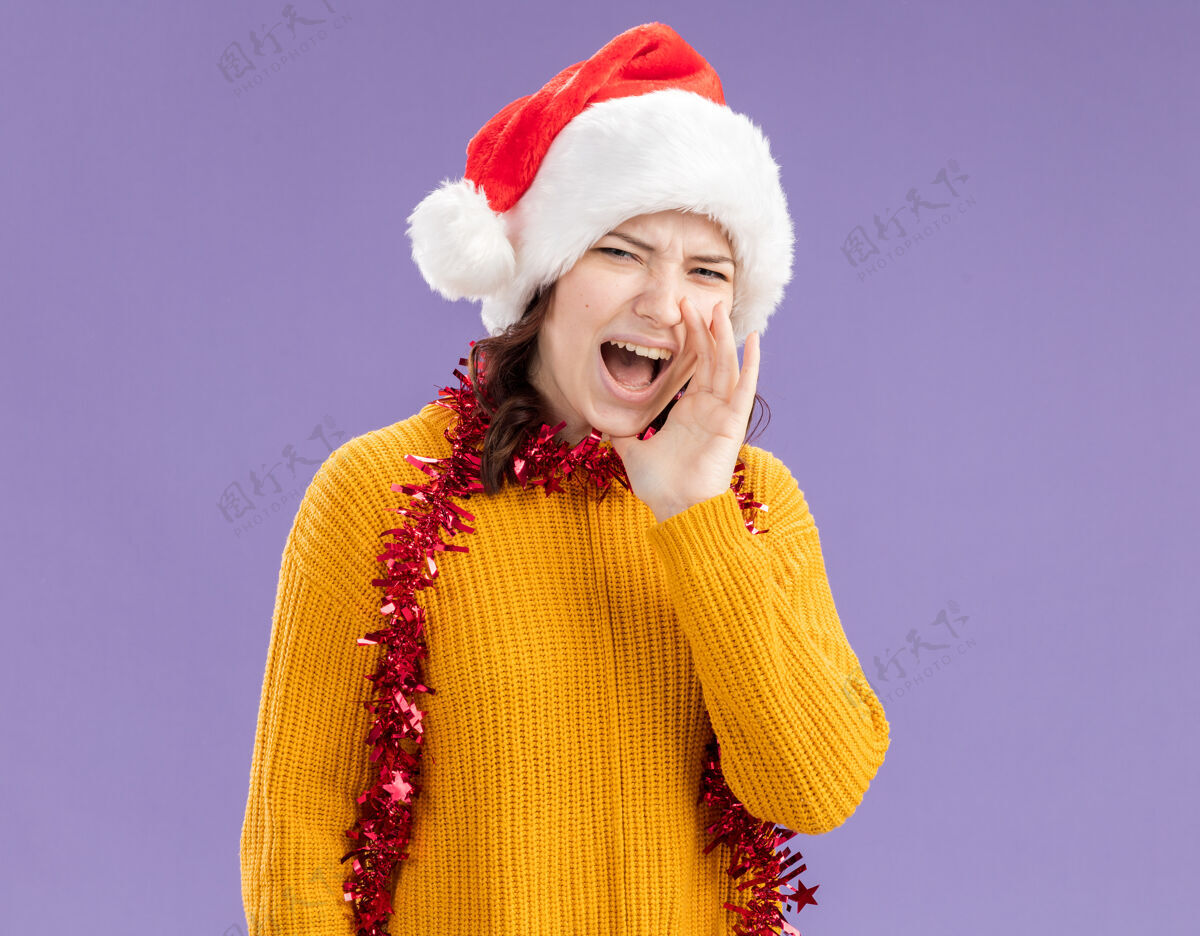 有人戴着圣诞帽 脖子上戴着花环的恼怒的斯拉夫女孩 手紧握着嘴 对着一个看着被隔离在紫色背景下 有复印空间的摄像机的人大喊大叫恼怒圣诞老人花环