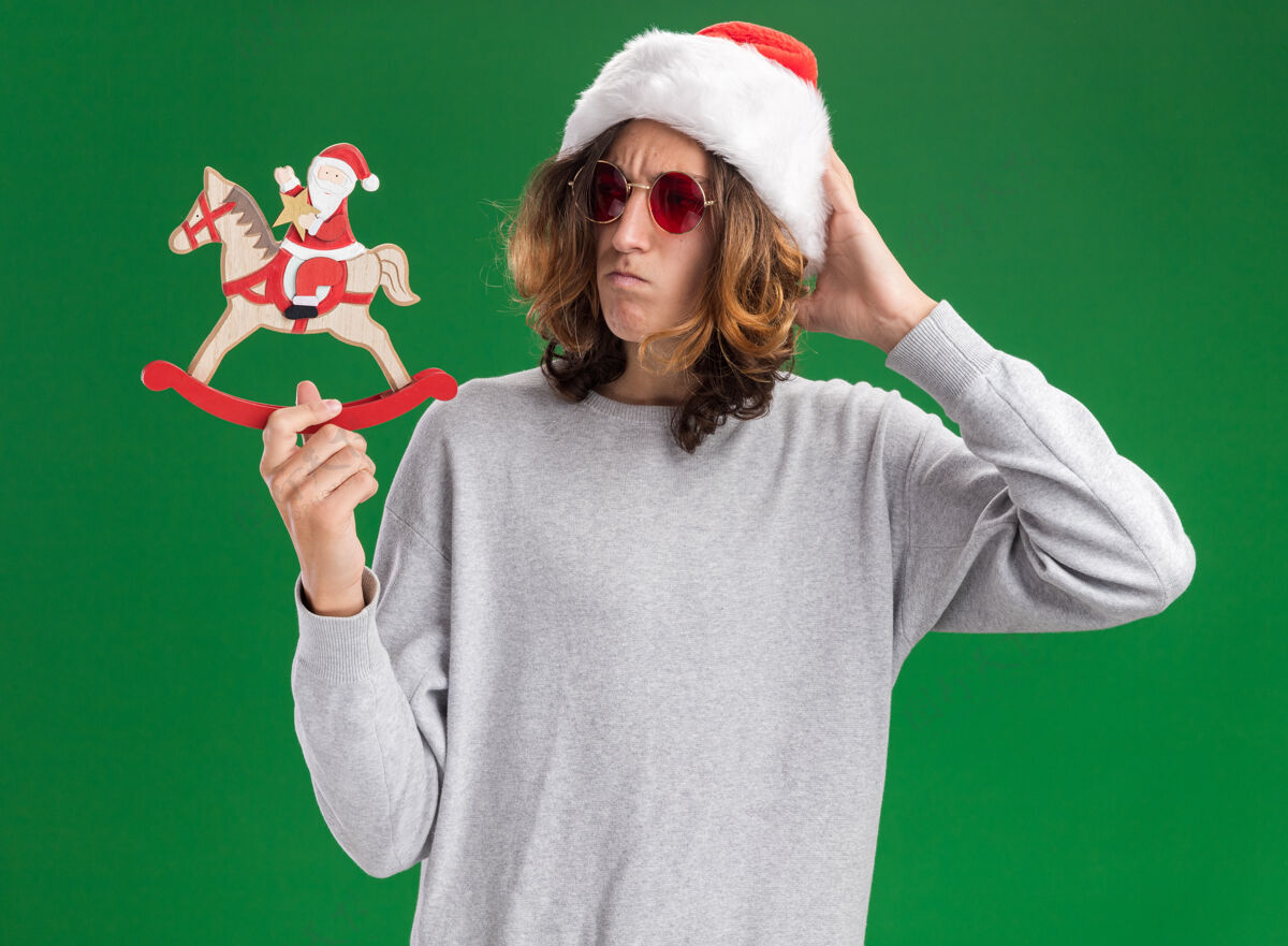 站着年轻人戴着圣诞老人帽 戴着红眼镜 手里拿着圣诞玩具 困惑地看着它 手放在头上 站在绿色的背景上年轻帽子圣诞节