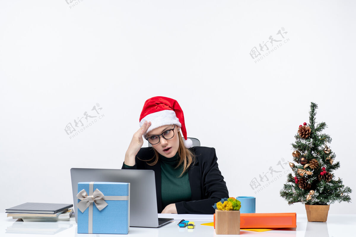 商务带着圣诞老人帽子的女商人坐在一张桌子旁 桌上放着圣诞树和礼物坐着圣诞老人女人