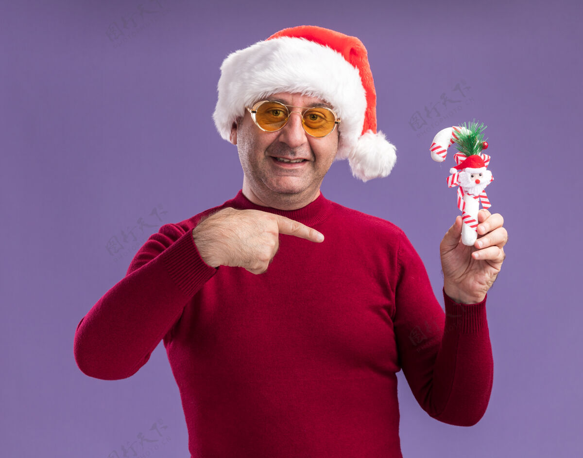 微笑中年男子戴着圣诞老人帽 戴着黄色眼镜 手里拿着圣诞糖果手杖 用食指指着它 站在紫色的背景上微笑着手杖帽子中间