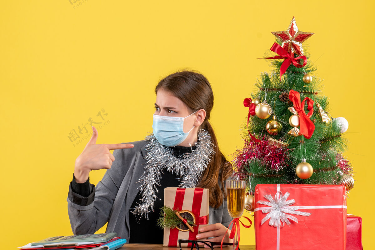 年轻前视图戴着医用面具的年轻女孩看着左边的圣诞树和礼物鸡尾酒成人面具桌子