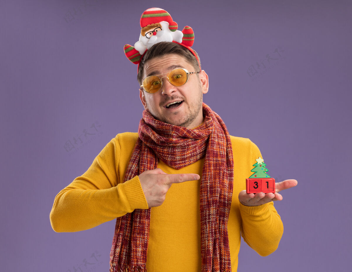 立方体快乐的年轻人穿着黄色高领毛衣 戴着暖和的围巾和眼镜 戴着滑稽的帽沿 头上戴着圣诞老人 在紫色的墙上展示着玩具立方体和新年的日期高领毛衣边缘围巾