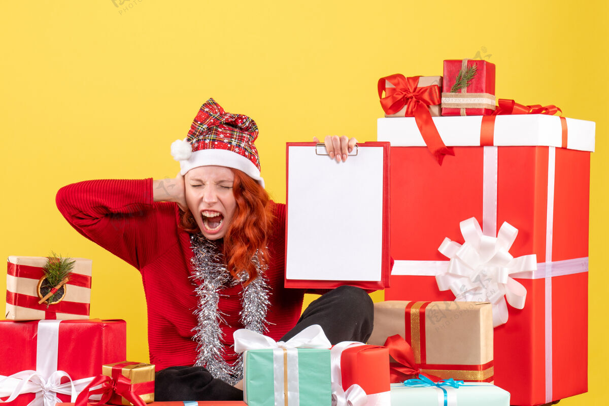 礼物坐在圣诞礼物旁边的年轻女子的正面图 黄色墙上有一张文件纸条年轻女性节日购物