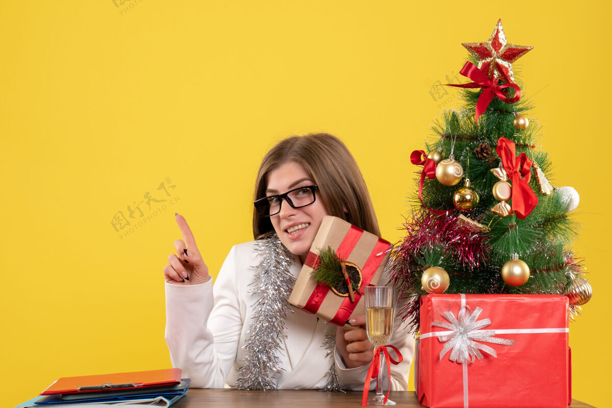 人正面图：女医生坐在桌子前 手里拿着黄色背景的礼物 还有圣诞树和礼品盒健康成人女医生