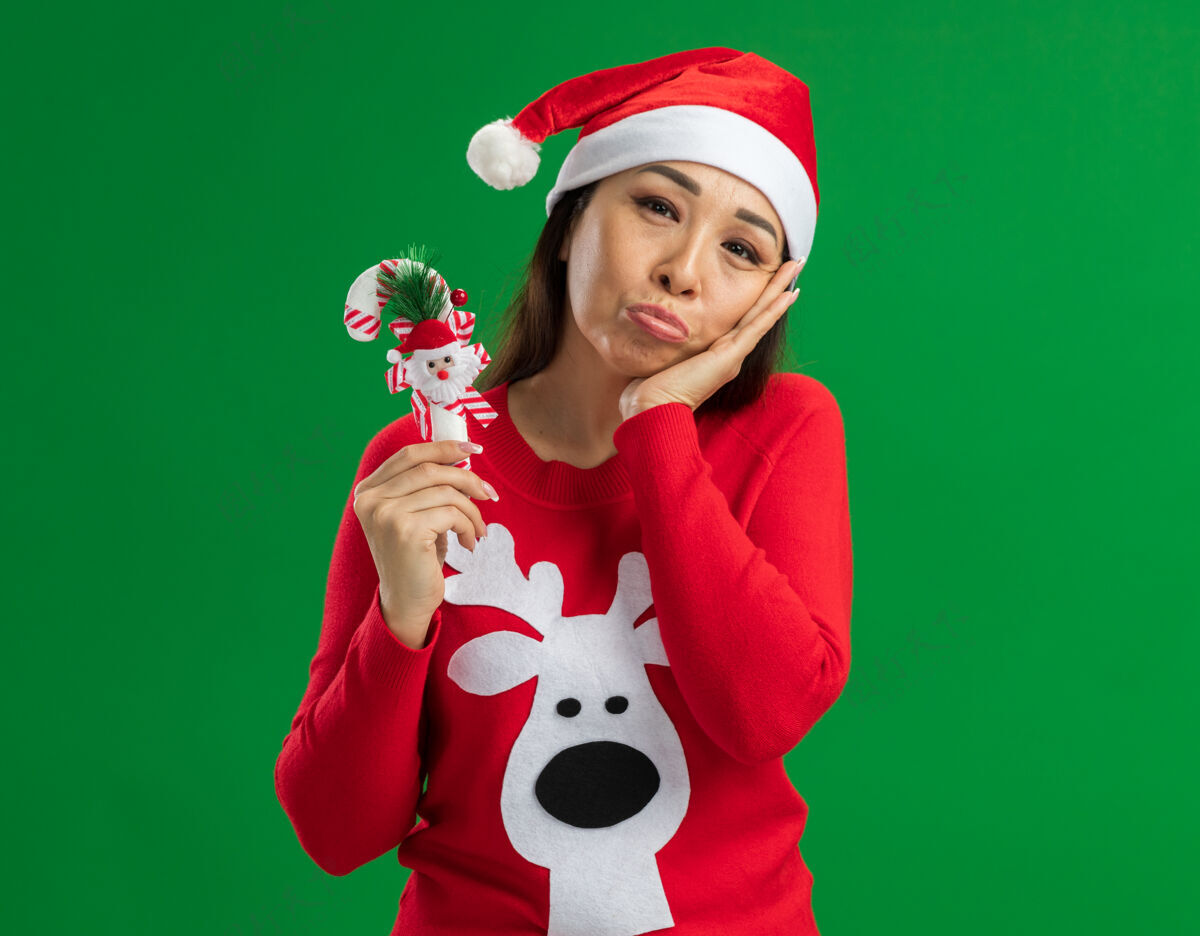 抱着戴着圣诞老人帽 穿着红色毛衣 手持圣诞糖果手杖的年轻女子站在绿色背景下 带着悲伤的表情看着镜头表情圣诞节悲伤