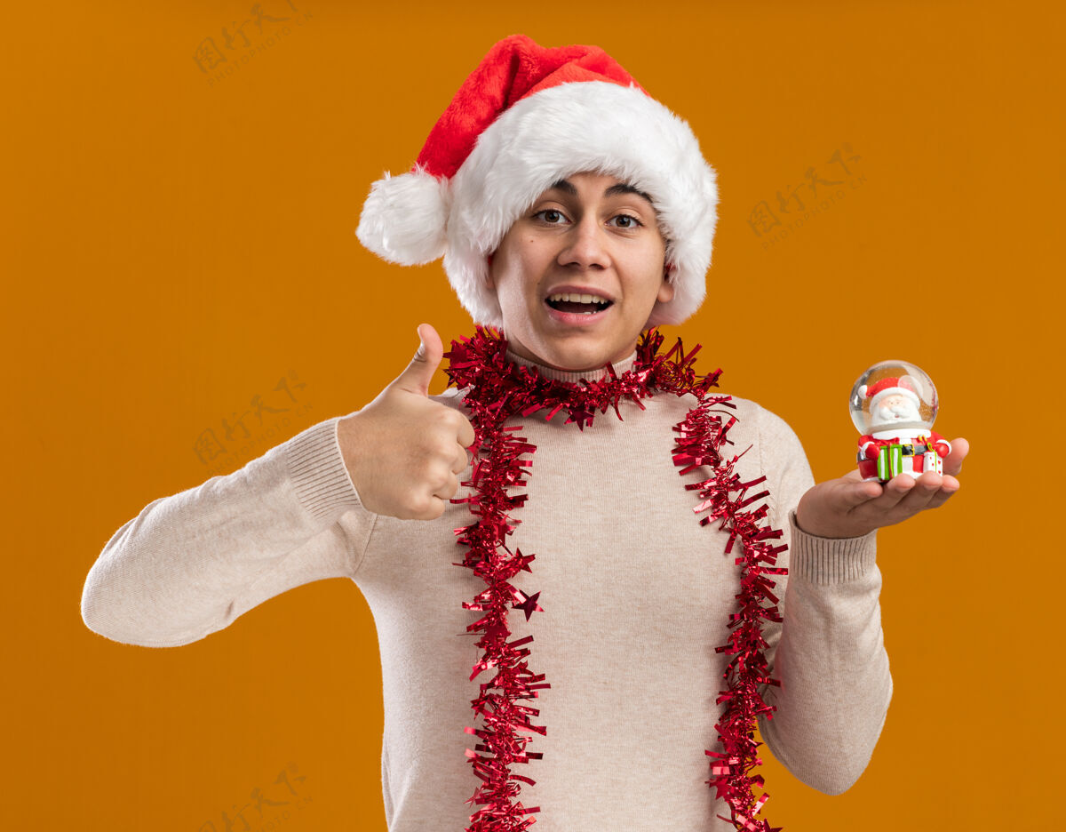 圣诞节微笑的年轻人戴着圣诞帽 脖子上戴着花环 手里拿着圣诞玩具 在黄色的墙上孤立地竖起大拇指展示帽子拇指