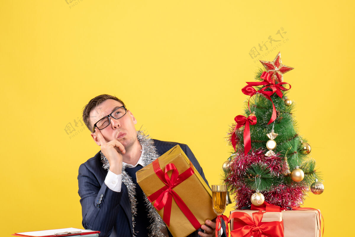西装前视图困惑的人戴着眼镜坐在圣诞树附近的桌子上 黄色的礼物男性困惑圣诞节