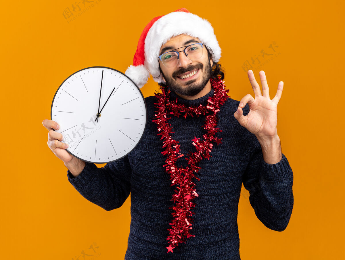 男人微笑的年轻帅哥戴着圣诞帽 脖子上戴着花环 手持挂钟 在橙色背景上显示出良好的姿态墙壁年轻展示