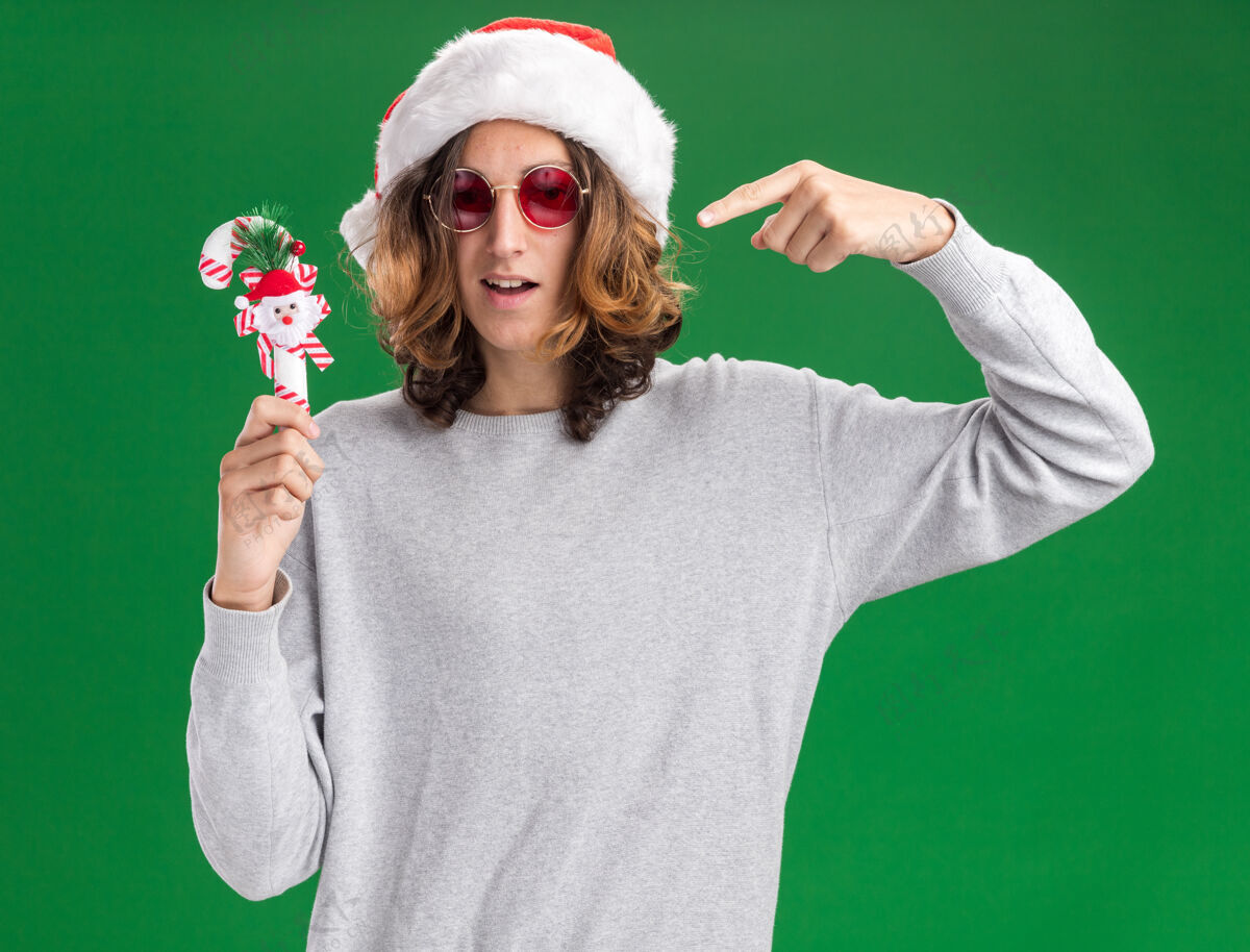 高兴戴着圣诞老人帽 戴着红眼镜 手持圣诞糖果手杖的年轻人微笑着用食指指着站在绿色背景上的手杖站着快乐帽子