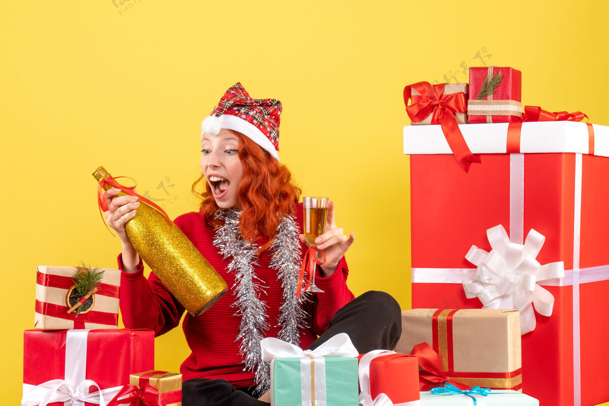 前面年轻女子围坐在圣诞礼物旁 在黄色墙壁上用香槟庆祝的正面视图快乐圣诞节年轻