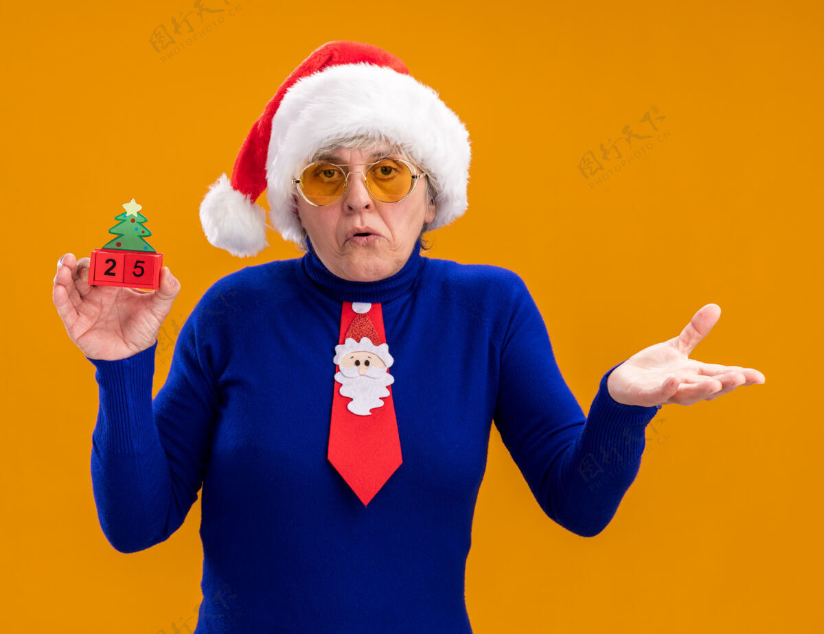 老人戴着太阳眼镜 戴着圣诞帽 系着圣诞领带 手拿圣诞树饰物的困惑的老妇人 在橙色背景上保持手的张开与复制空间隔离装饰品圣诞老人橙色