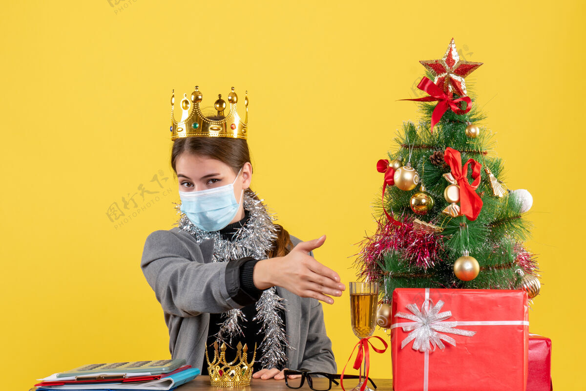 穿着正面图戴着医用面罩戴着皇冠的年轻女孩手拿圣诞树和鸡尾酒礼物年轻手