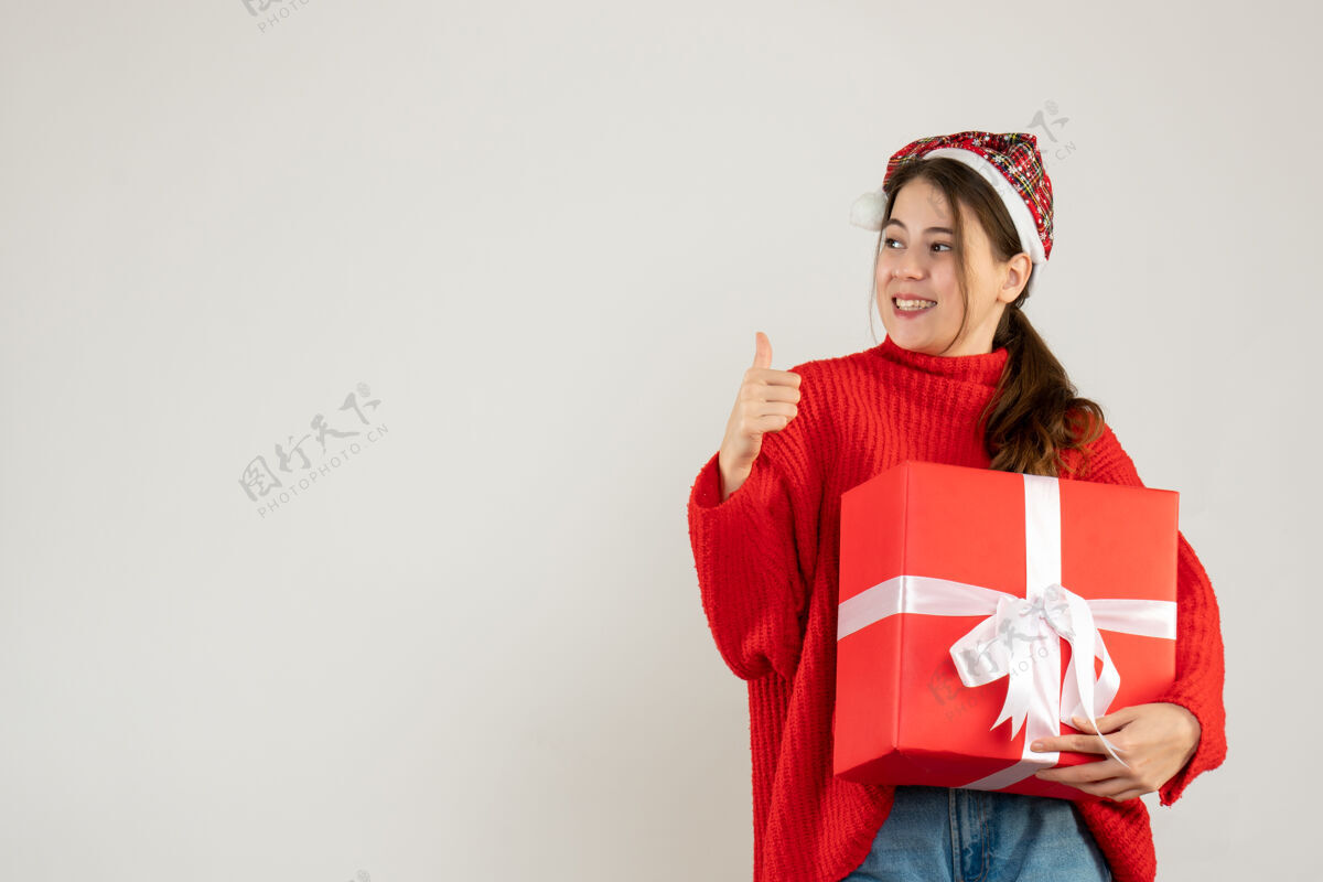 圣诞节戴着圣诞帽的可爱女孩微笑着拿着礼物站在白色的地板上竖起大拇指可爱漂亮礼物