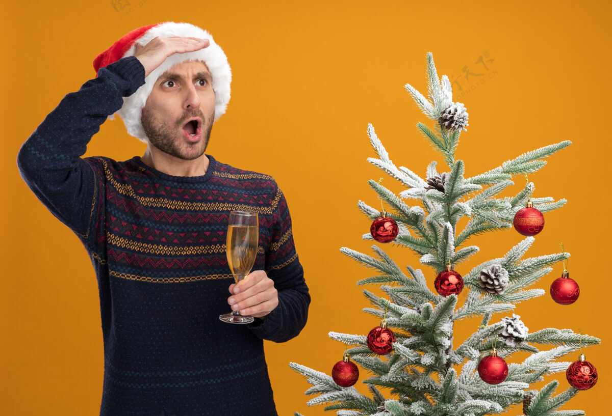 距离令人印象深刻的年轻白人男子戴着圣诞帽站在装饰圣诞树附近拿着一杯香槟望着远方隔离在橙色墙上近侧面印象深刻