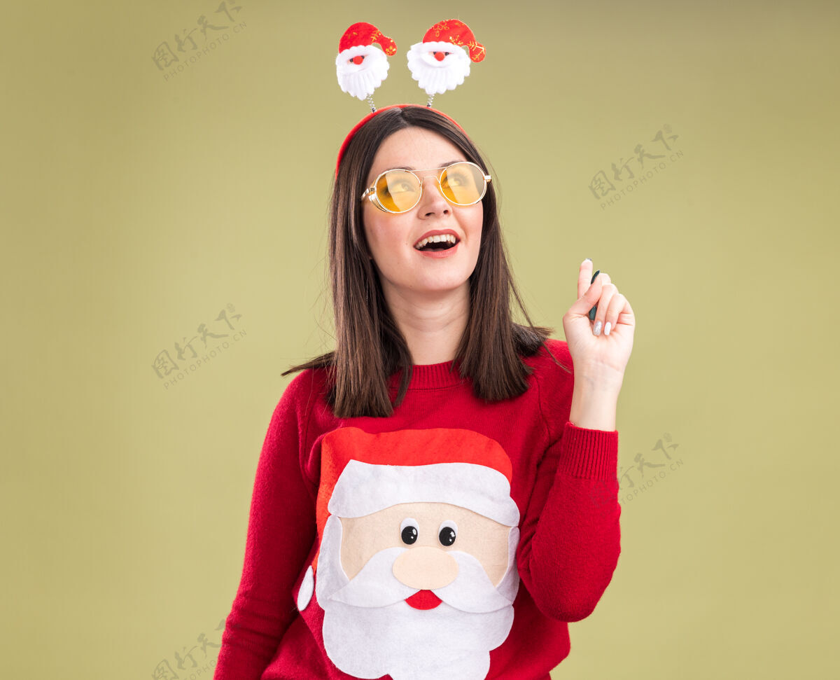 指向令人印象深刻的年轻漂亮的白人女孩穿着圣诞老人毛衣和头带眼镜看着和指着橄榄绿背景与复制空间隔离女孩印象深刻漂亮