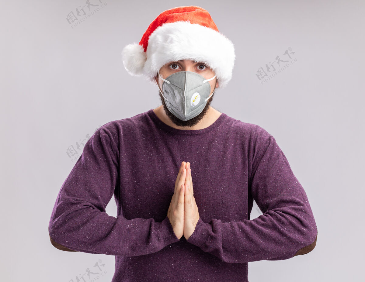 站着身穿紫色毛衣 头戴圣诞老人帽 戴着护面面具的年轻人手拉着手 像在祈祷似的 站在白色背景下焦急地看着镜头保护在一起担心