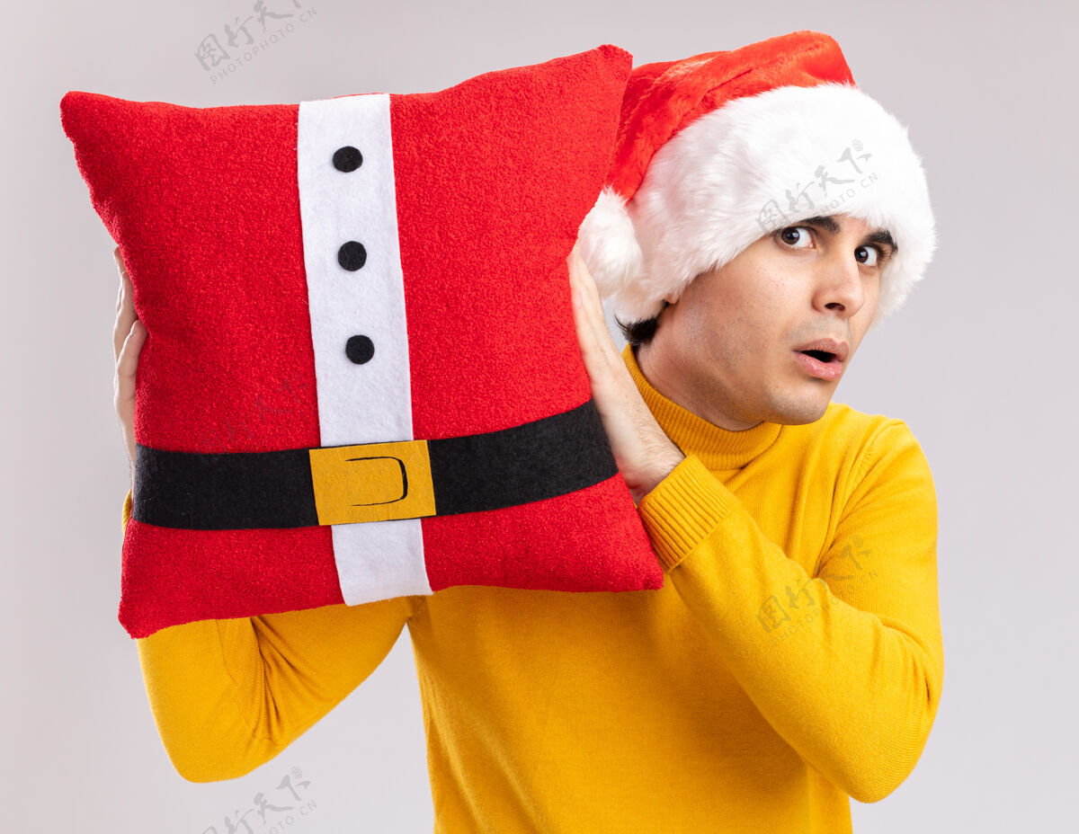 站着穿着黄色高领毛衣 戴圣诞帽的年轻人拿着圣诞枕头站在白色背景下惊讶地看着相机年轻圣诞节帽子