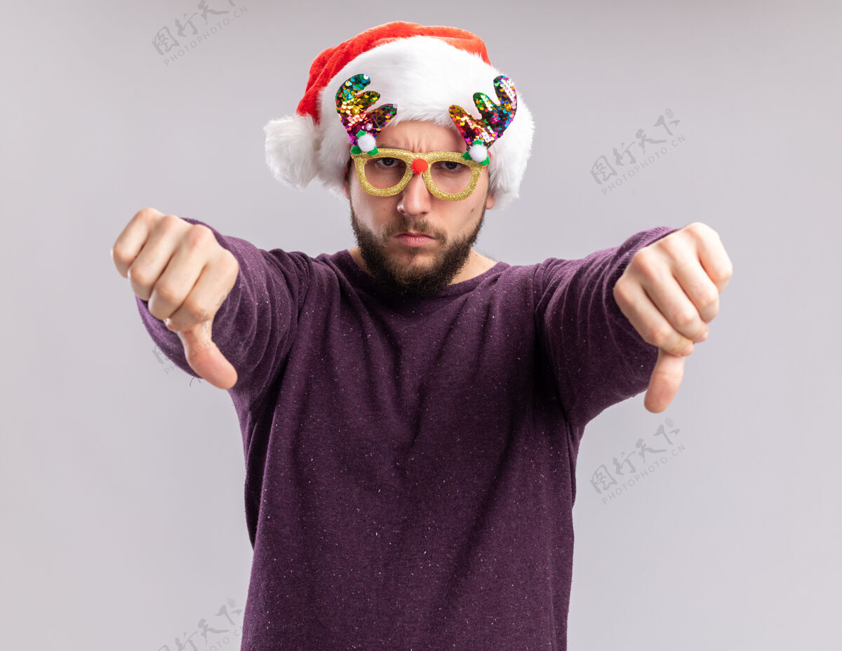 脸一个穿着紫色毛衣 戴着圣诞帽 戴着滑稽眼镜的年轻人站在白色背景上 怒气冲冲地看着镜头 大拇指朝下眼镜毛衣站着