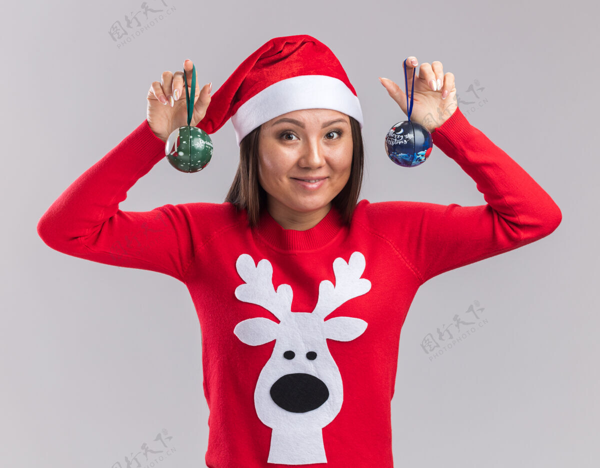 女孩笑容满面的亚洲女孩戴着圣诞帽 穿着毛衣 手里拿着圣诞树上的球 背景是白色的举行树帽子