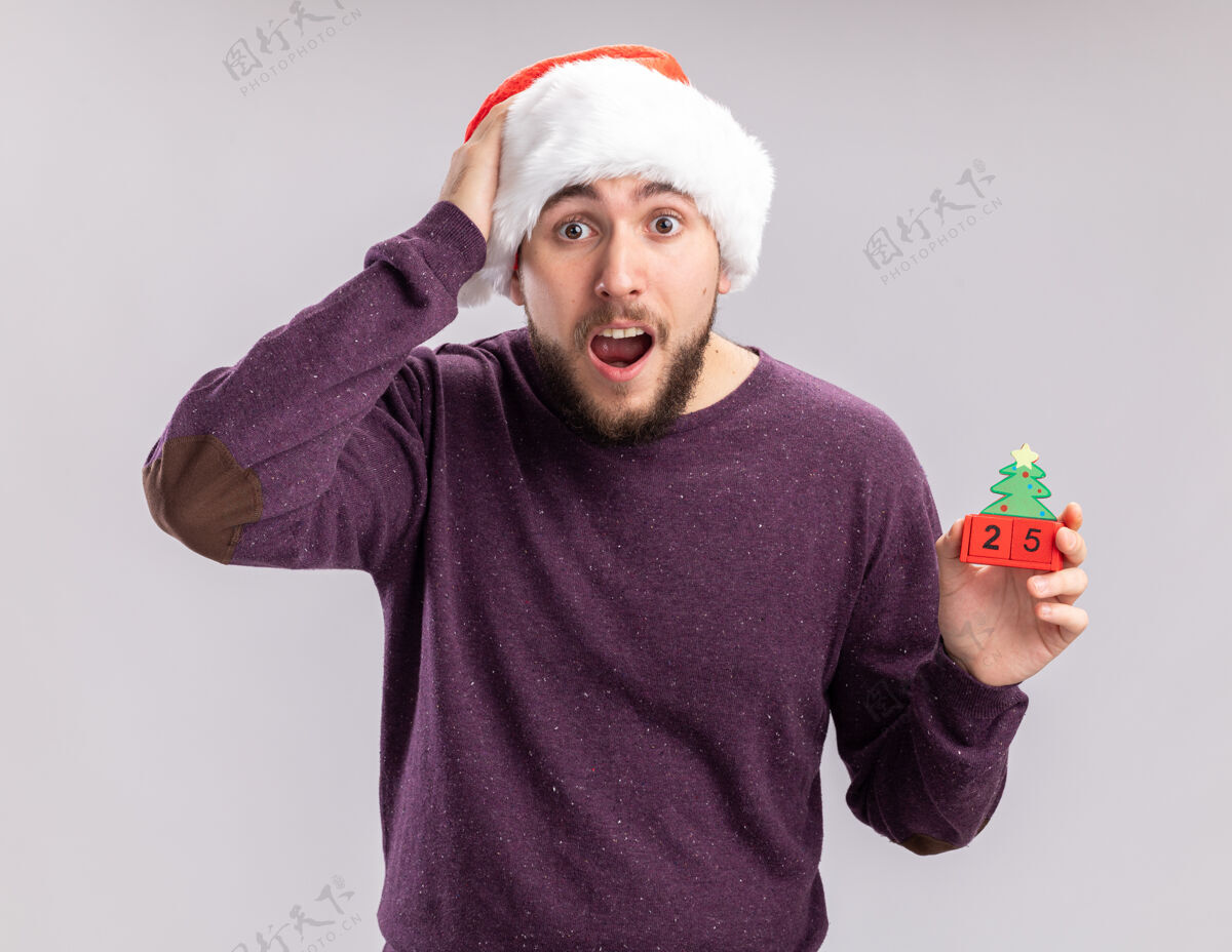 立方体身穿紫色毛衣 戴圣诞帽的年轻人站在白色背景下 展示着新年约会的立方体 看着镜头 惊讶不已站着圣诞老人毛衣
