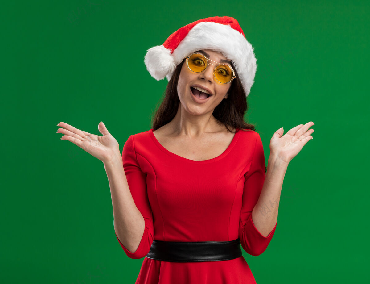 眼镜兴奋的年轻漂亮女孩戴着圣诞帽和眼镜 两手空空地看着隔离在绿色背景上的相机女孩兴奋空虚