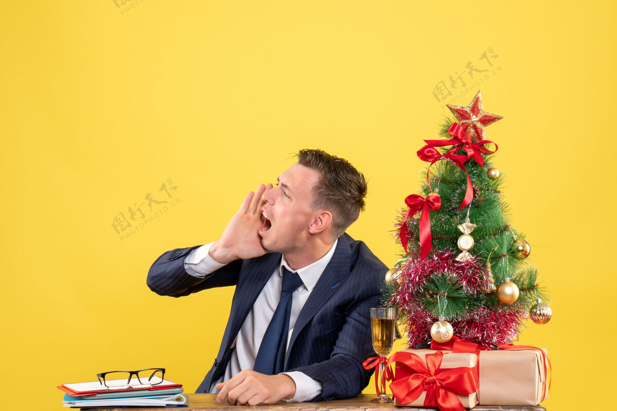 办公室愤怒的男人打电话给坐在圣诞树旁桌子旁的人 黄色墙上挂着礼物某人商务愤怒