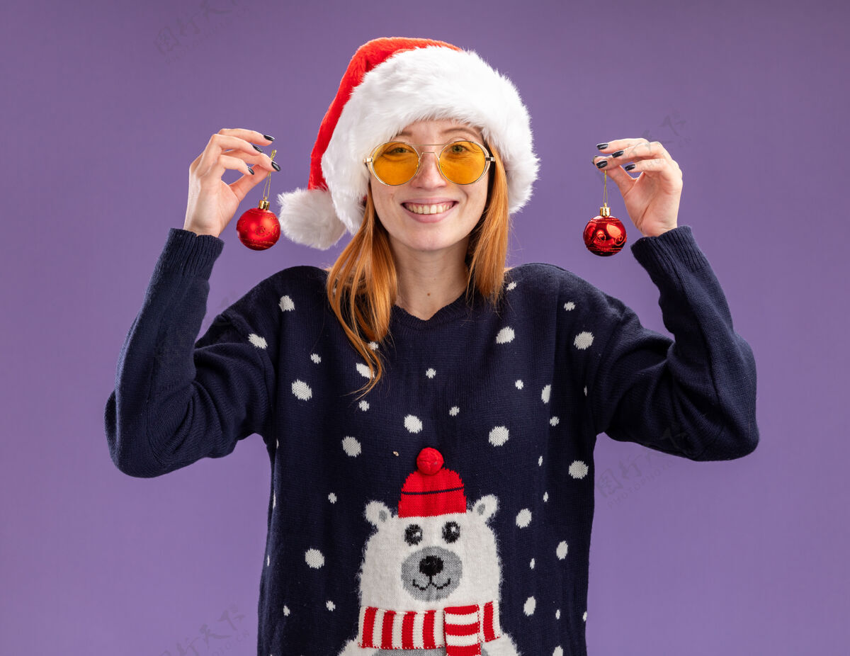 年轻人微笑着的年轻漂亮女孩穿着圣诞毛衣 戴着圣诞帽 戴着一副眼镜 手里拿着圣诞球 背景是紫色的女孩抱着微笑