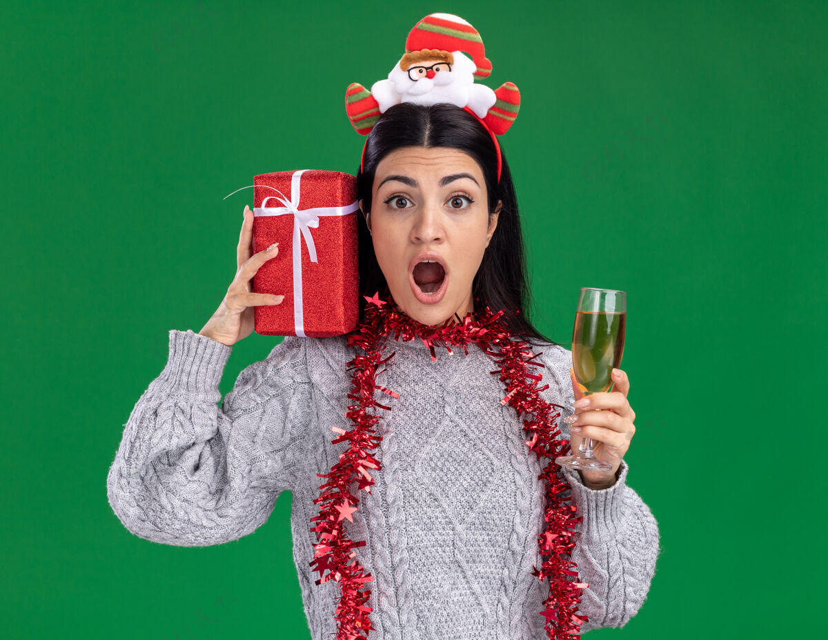 肩膀年轻的白人女孩戴着圣诞老人的头带 脖子上戴着金箔花环 肩上扛着礼包 绿色的墙上隔着一杯香槟脖子玻璃头带