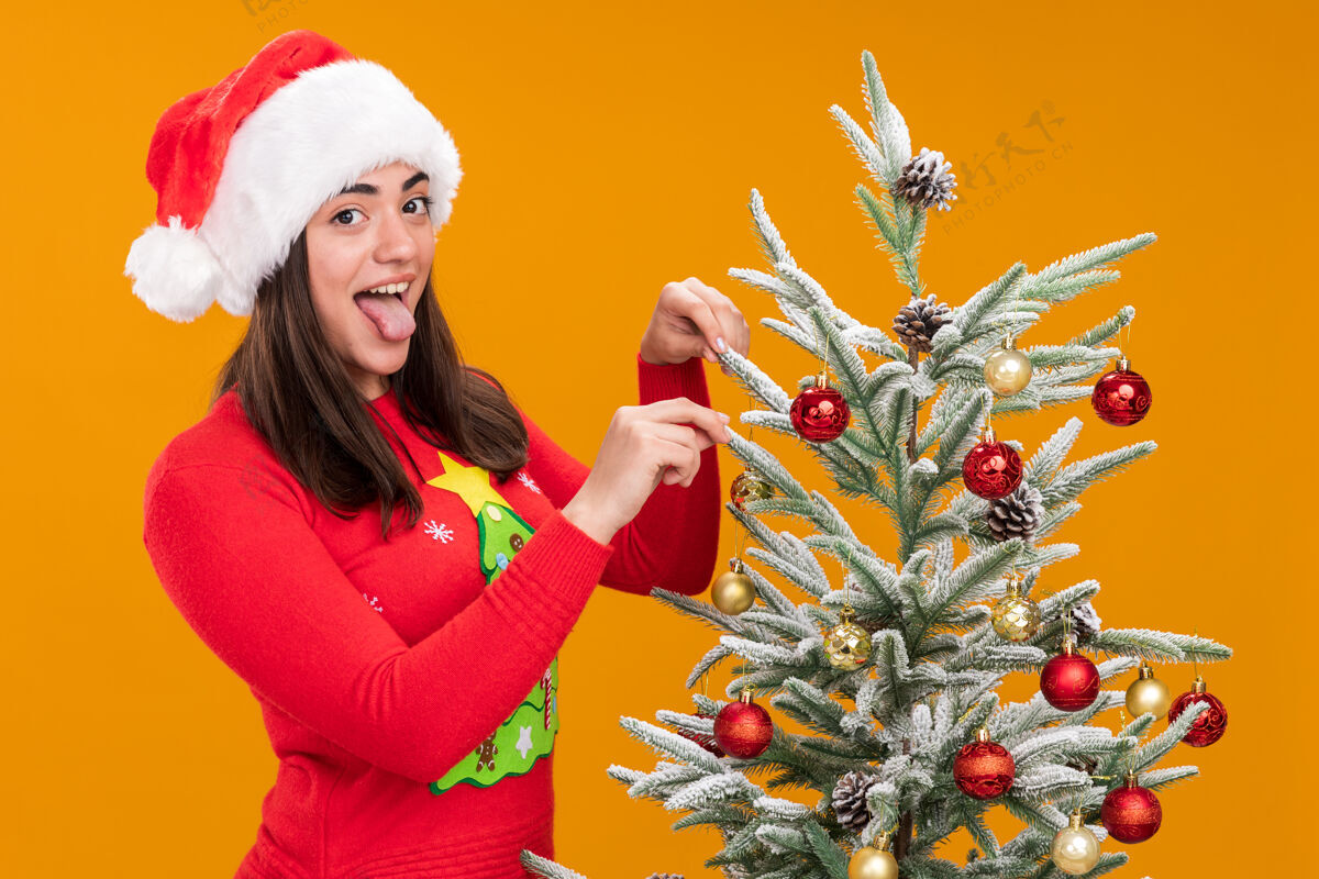 圣诞树快乐的年轻白人女孩戴着圣诞帽 伸出舌头站在圣诞树旁 装饰着橙色背景的玩具 还有复制空间圣诞老人站着橙色
