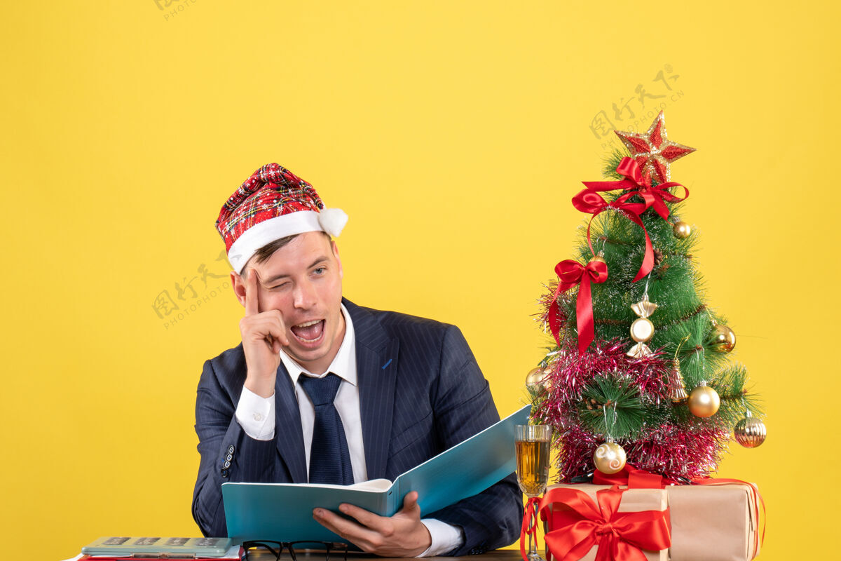 眼睛商人眨着眼睛坐在圣诞树旁的桌子前 黄色的墙上摆着礼物商人礼物前面