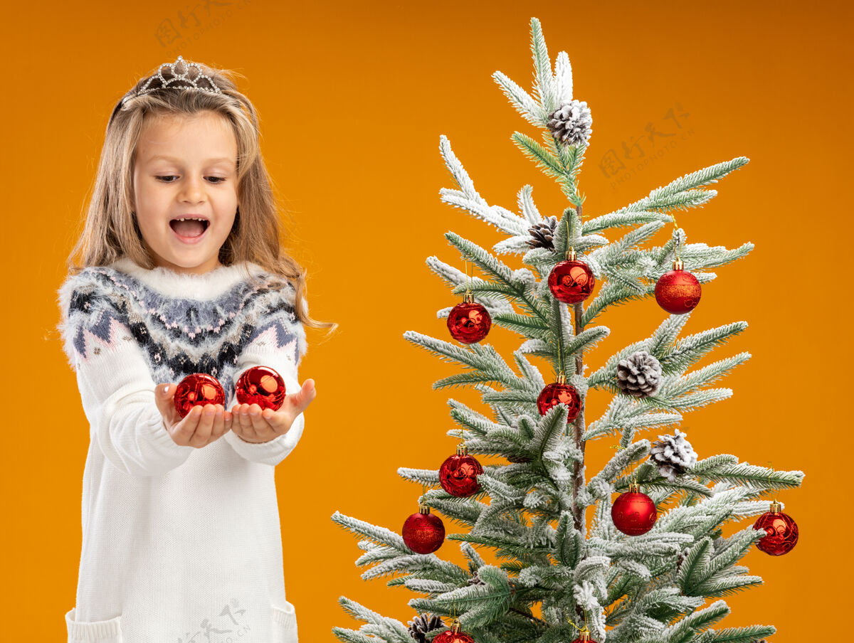站着快乐的小女孩站在圣诞树旁 戴着头饰 脖子上戴着花环 手里拿着孤立在橙色墙上的圣诞球快乐脖子附近