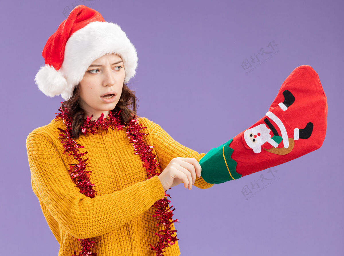 年轻惊讶的年轻斯拉夫女孩戴着圣诞帽 脖子上戴着花环 把她的手插在圣诞长袜里 在紫色背景上 留有复制空间圣诞节脖子花环