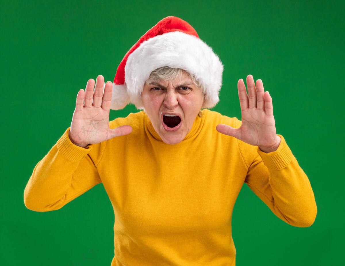 恼怒戴着圣诞老人帽子的老妇人恼怒地对着一个人大喊大叫 她正看着被隔离在紫色背景上的相机 还有复印空间有人老人帽子