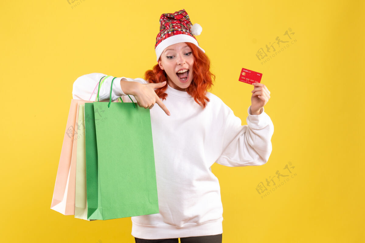 圣诞节黄墙上拿着购物包和银行卡的年轻女子的正视图购物微笑包裹