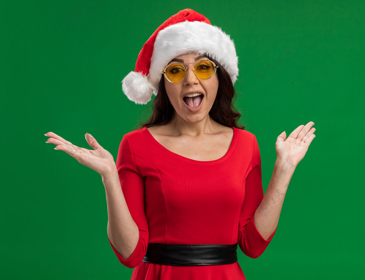 手年轻漂亮的女孩戴着圣诞帽 戴着眼镜 两手空空地看着隔离在绿色背景上的相机帽子年轻空虚