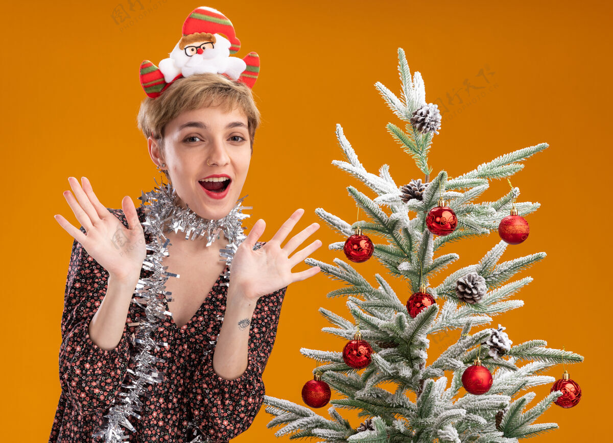 站着年轻漂亮的女孩戴着圣诞老人的头带 脖子上戴着金属丝的花环 站在装饰过的圣诞树旁 两手空空的 孤零零地站在橙色的墙上印象深刻圣诞节显示