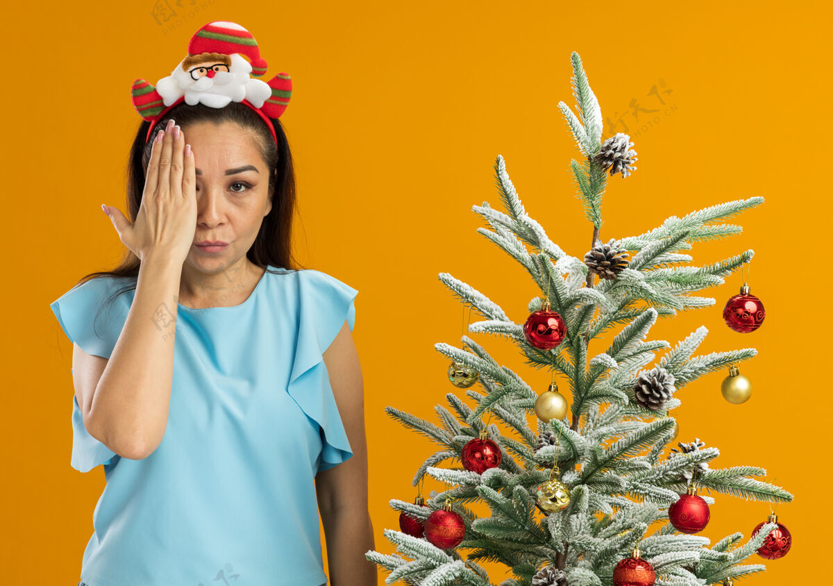 边缘身穿蓝色上衣的年轻女子头戴滑稽的圣诞戒指 严肃的脸遮住一只眼睛 手站在橙色背景下的圣诞树旁 看着摄像机圣诞节树站立