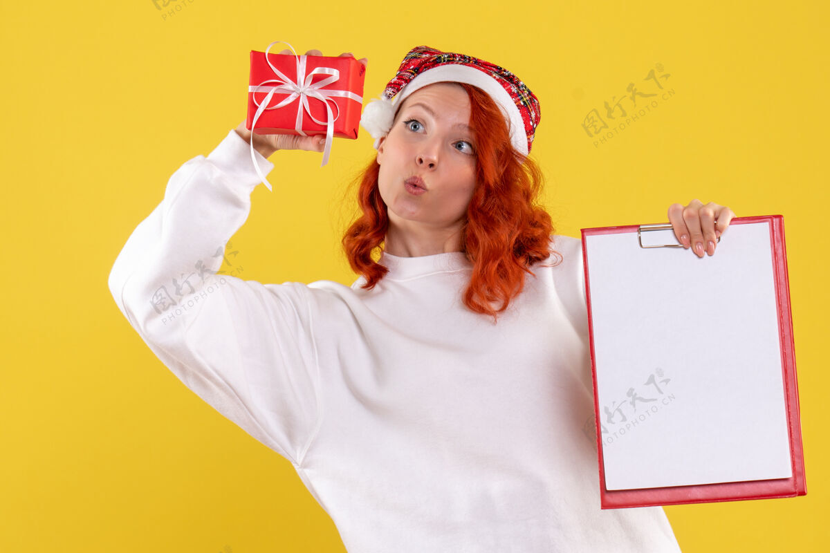 礼物手持小圣诞礼物和黄色墙上的文件笔记的年轻女子的正面视图节日礼物圣诞节
