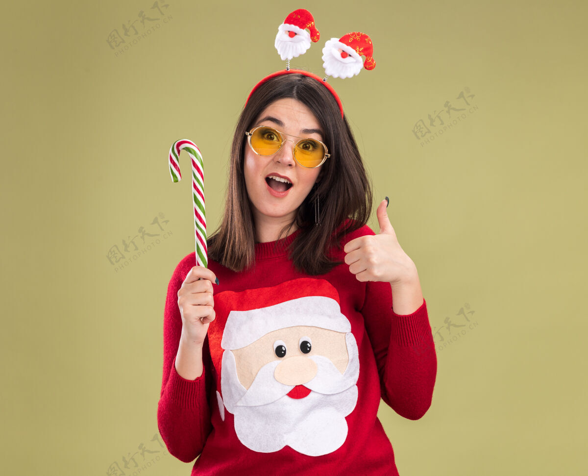 圣诞快乐令人印象深刻的年轻漂亮的白人女孩穿着圣诞老人毛衣和头带眼镜举行传统的圣诞糖果手杖漂亮眼镜年