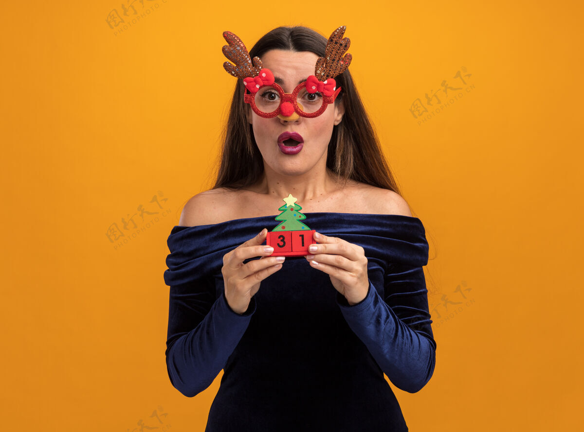 圣诞节穿着蓝色连衣裙 戴着圣诞眼镜 拿着橙色背景上孤立的玩具的年轻漂亮女孩惊讶不已年轻惊讶眼镜