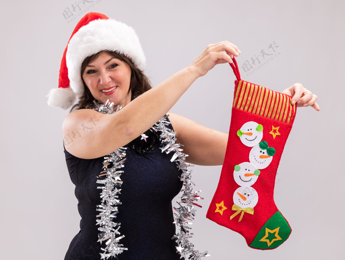 微笑微笑的中年妇女戴着圣诞帽 脖子上戴着金属丝花环 手里拿着圣诞长袜 看着隔离在白色背景上的相机周围金属丝中年