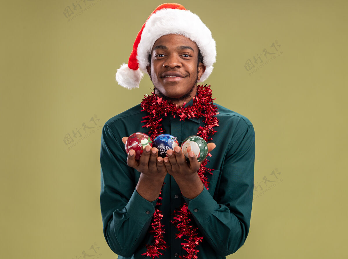圣诞节戴着圣诞帽 戴着花环 拿着圣诞球的非裔美国人站在绿色背景下 面带微笑地看着摄像机帽子站着球
