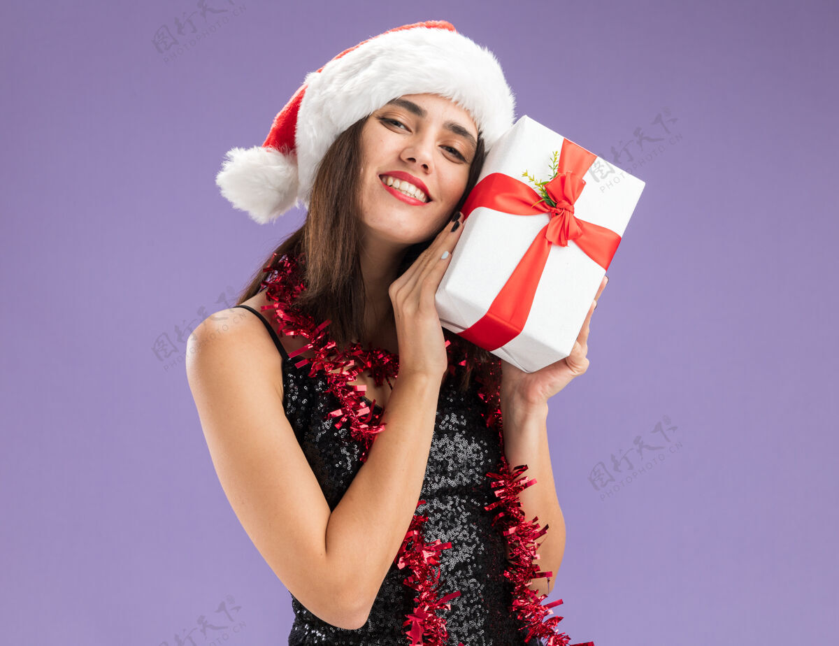 帽子可爱的年轻漂亮女孩戴着圣诞帽 脖子上戴着花环 脸上围着礼品盒 背景是紫色的抱着女孩年轻