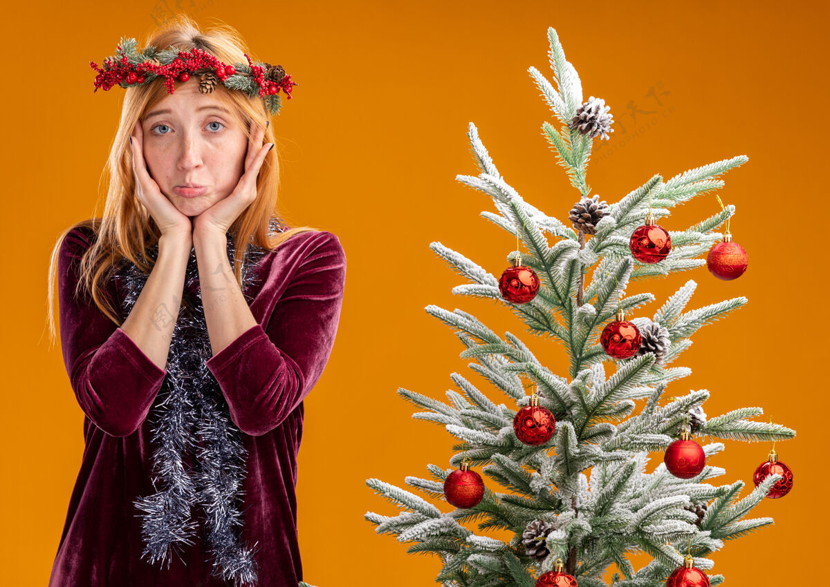 花环站在圣诞树旁的悲伤的年轻漂亮的女孩穿着红色的裙子 脖子上戴着花环 双手搂着孤立在橙色背景上的脸脖子圣诞节衣服