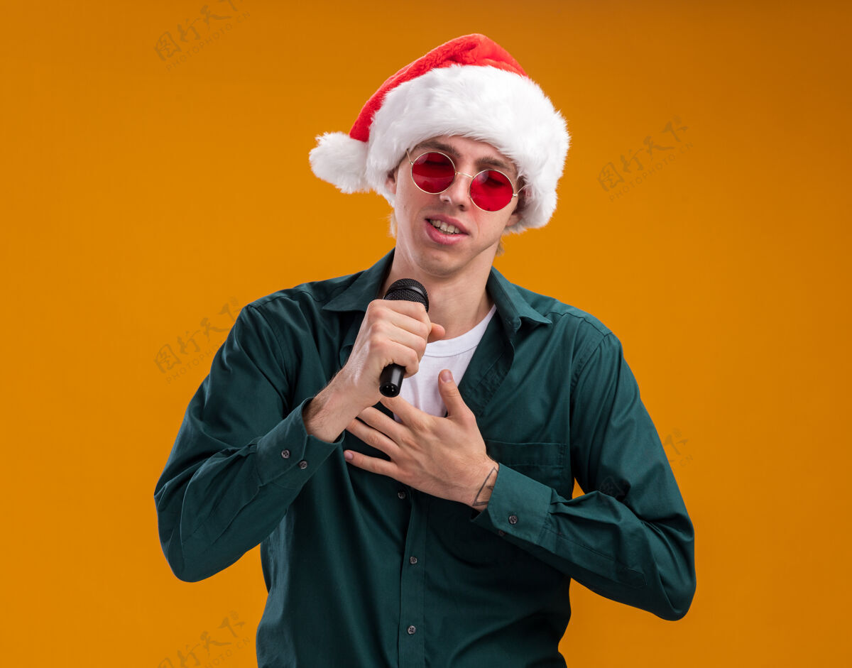 帽子戴着圣诞帽 戴着眼镜 手持麦克风 手放在胸前 闭着眼睛 在橙色背景下唱歌的年轻金发男人手麦克风圣诞老人