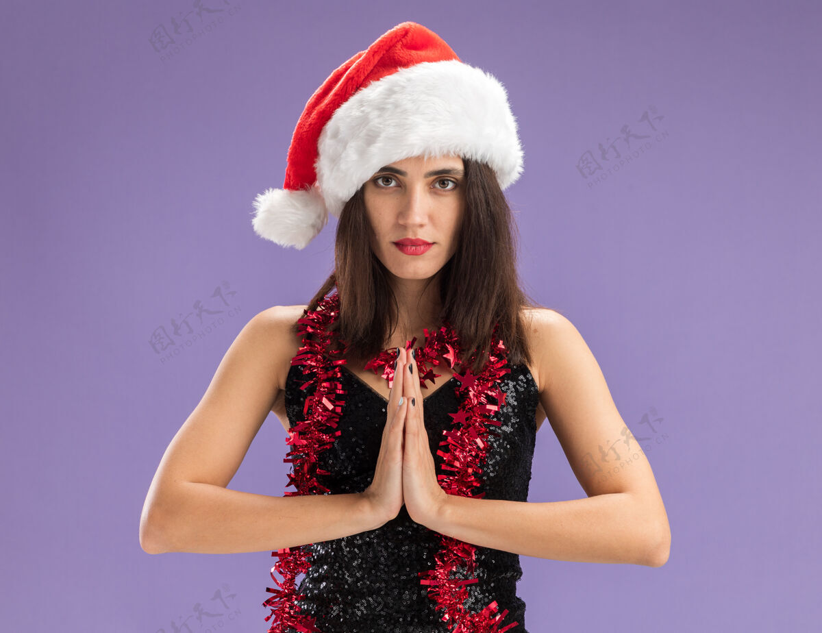 女孩年轻漂亮的女孩戴着圣诞帽 脖子上戴着花环 在紫色的背景上展示着与世隔绝的祈祷姿态帽子手势花环