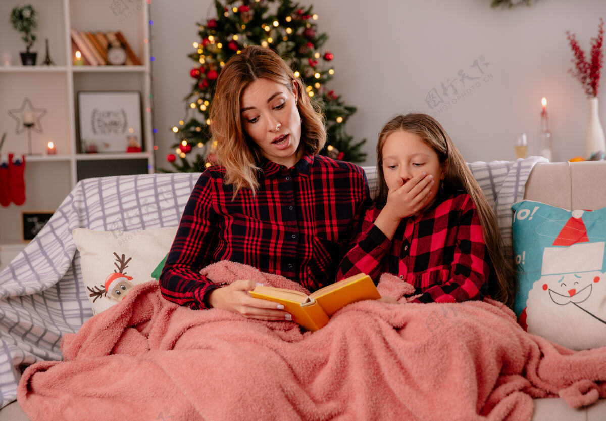女儿震惊的母女俩坐在沙发上读着铺满毯子的书 在家里享受圣诞节的时光坐时间享受