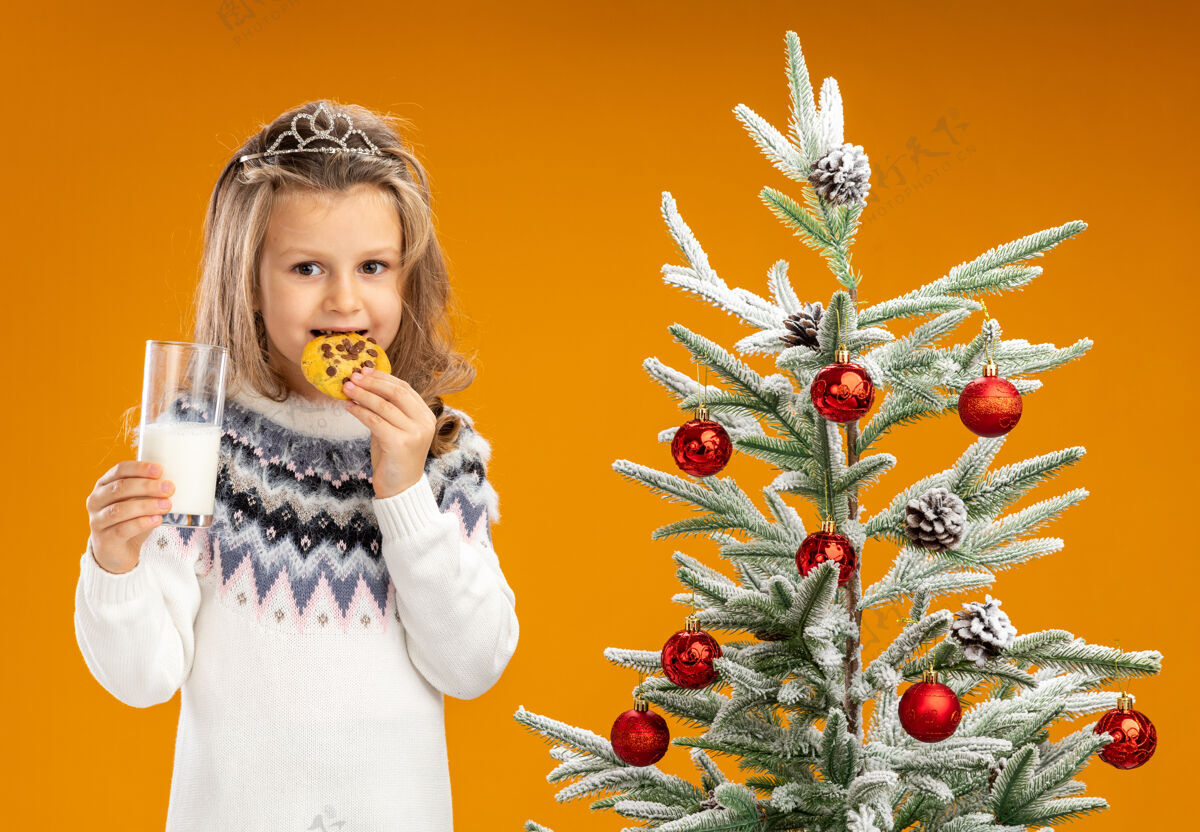 饼干高兴的小女孩站在圣诞树旁 戴着头饰 脖子上戴着花环 手里拿着一杯牛奶 品尝着橙色背景上孤立的饼干站着圣诞圣诞快乐
