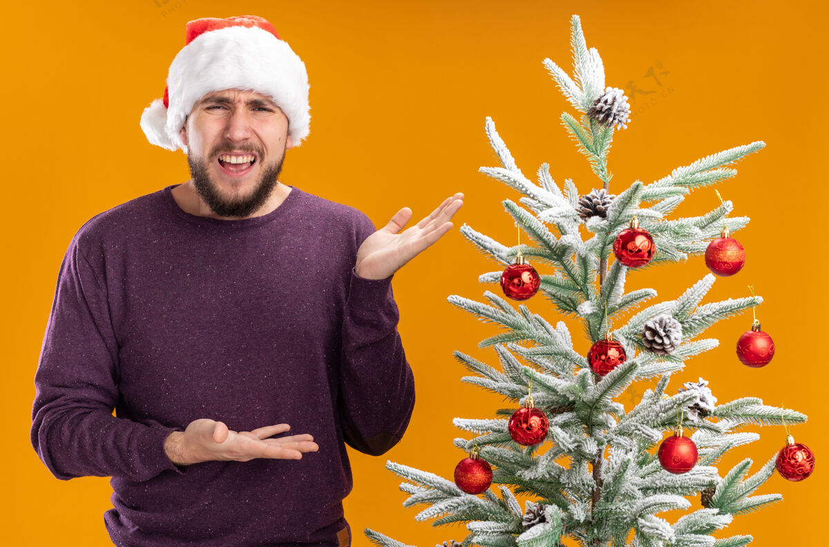 年轻身穿紫色毛衣 头戴圣诞帽的年轻人站在圣诞树旁 双手相拥 在橙色背景下显得困惑和不快站立手毛衣
